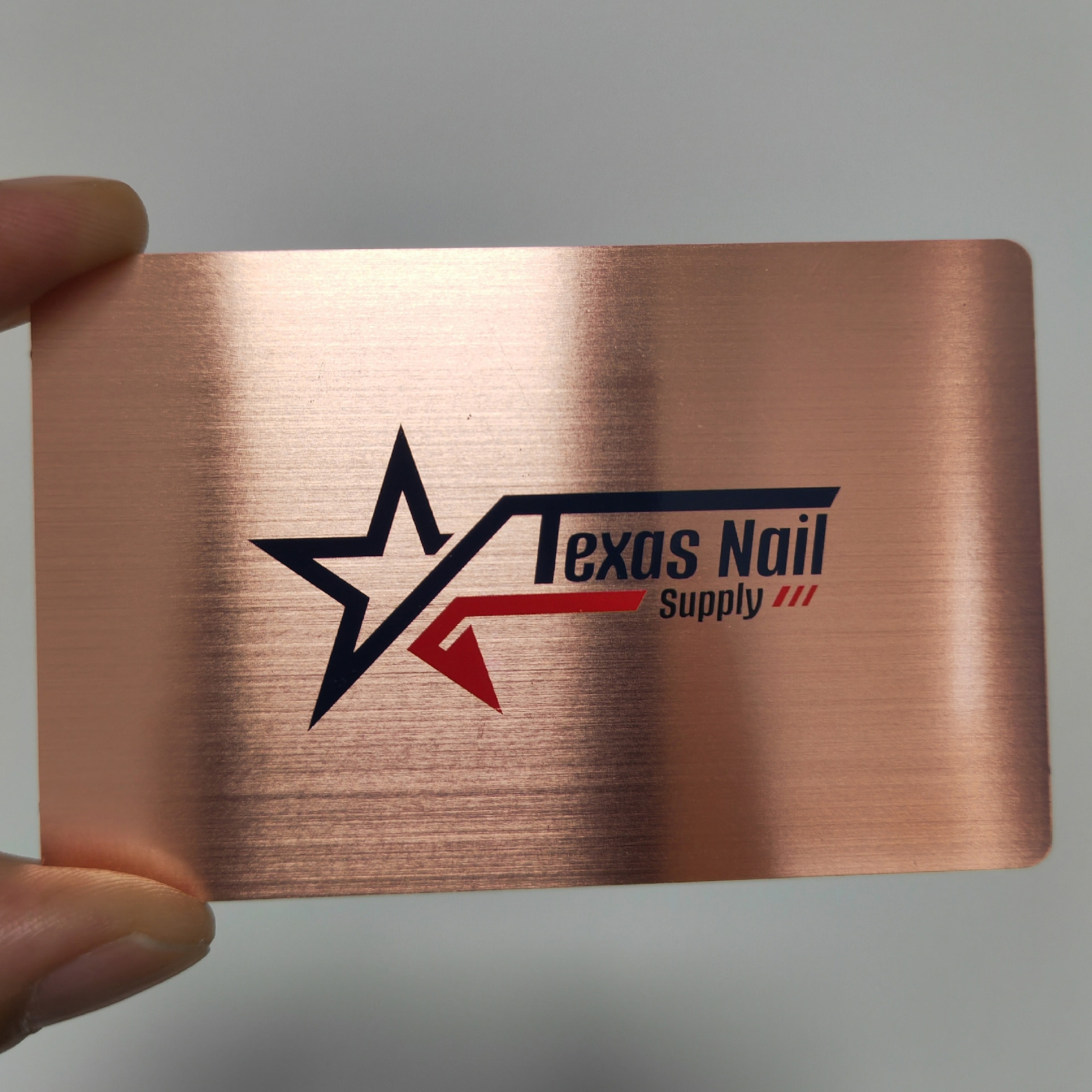 Una tarjeta de presentación de metal de cobre cepillado con logotipo Texas Nail Supply sostenida en la mano muestra la claridad de impresión sobre metal.