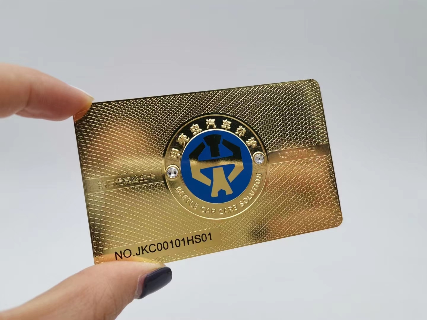 Una tarjeta de presentación de metal dorado texturizado con emblema redondo y pequeñas piedras destaca la elaboración prémium y el agarre.