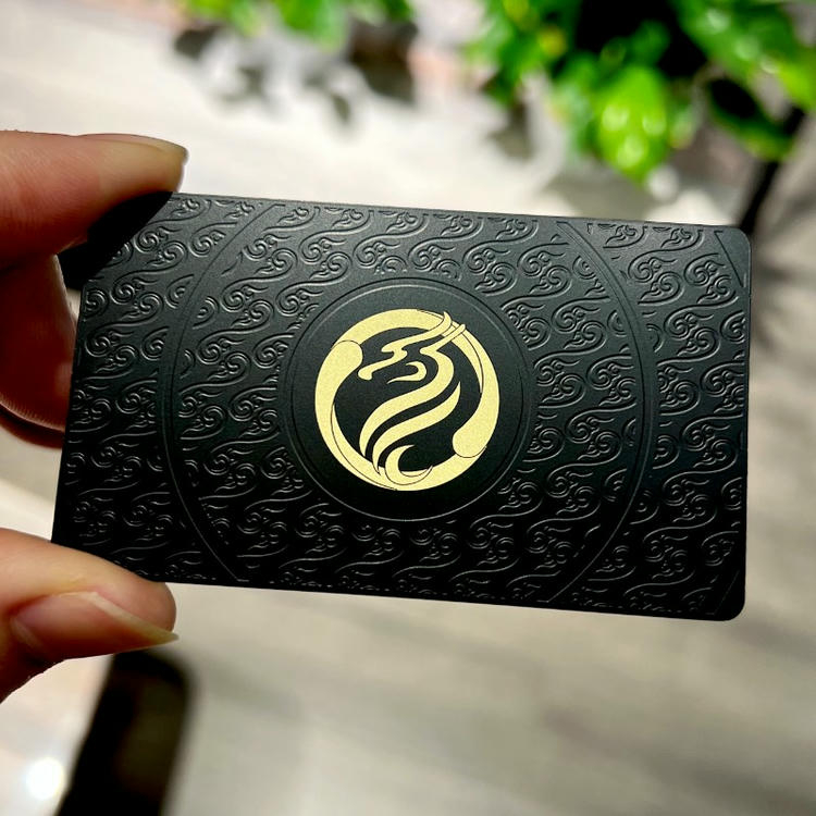 Esta tarjeta de visita metálica muestra una superficie negra texturizada con patrón en relieve y emblema dorado central.